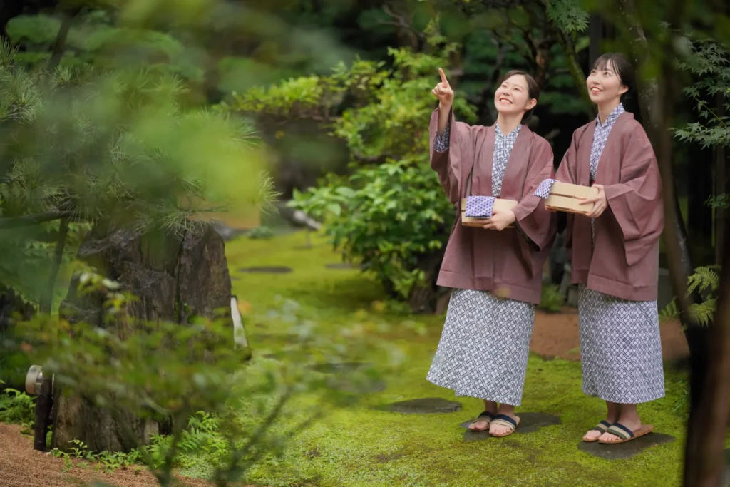 温泉の館内浴衣を着て、桶を持った二人の女性が庭で微笑んでいる様子
