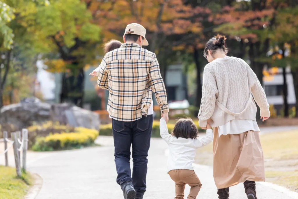 家族(夫婦と幼い子供2人)が手を繋いで紅葉の公園を歩いている。