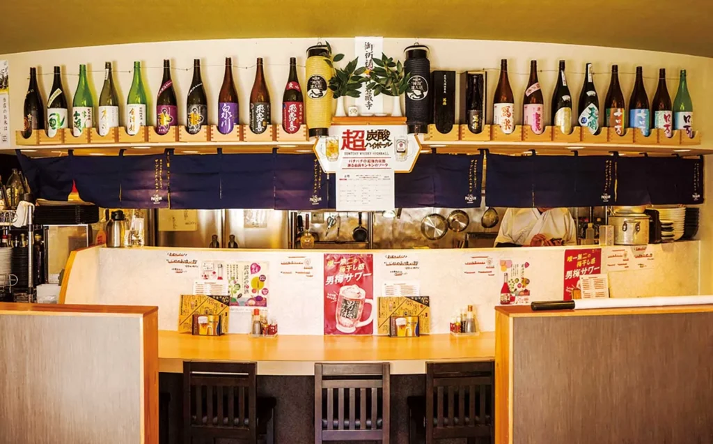居酒屋のカウンター席。頭上の棚に日本酒がずらりと並んでいる。
